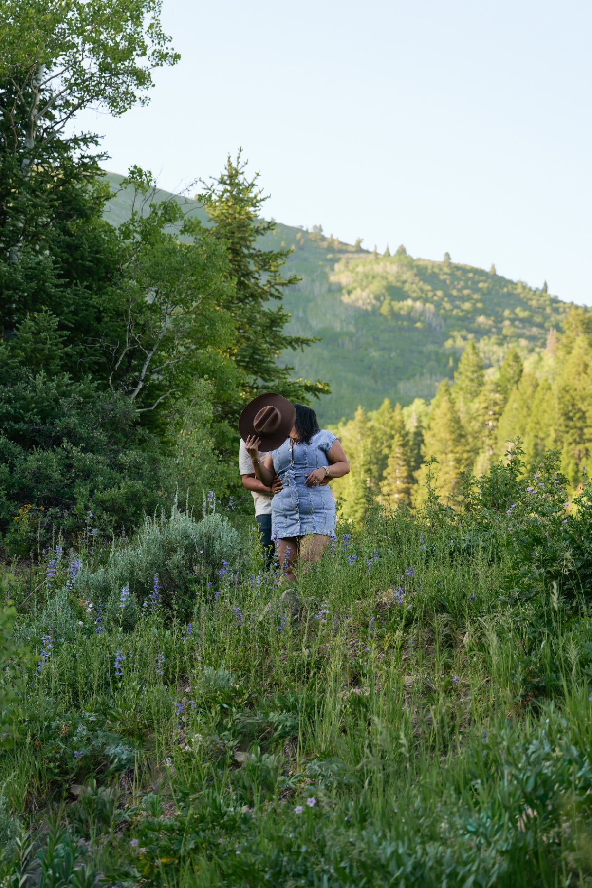 Couple kissing behind a brown hat in a mountain meadow